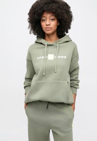 Olivovozelená mikina Nike s kapucňou na sťahovanie, predným vreckom a bielym logom. Kombinovaná s nohavicami na jogging v rovnakom odtieni s žltými akcentmi.