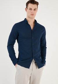Cămașă bleu marin cu mânecă lungă, cu guler cu nasturi, țesătură texturată și închidere cu nasturi pe față. Potrivită pentru ținute casuale sau semi-formale.