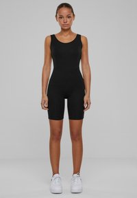 Černý bezrukávový body s těsným střihem a délkou šortek. Model nosí bílé tenisky. Hladká textura a minimalistický styl.