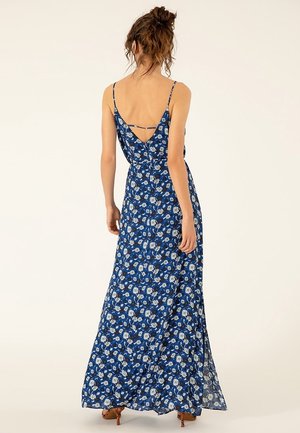 Femme aux cheveux bouclés attachés en un chignon lâche, portant une robe longue bleue à bretelles fines ornée de fleurs, avec un dos en V profond et des sandales à talons marron.