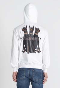Sudadera blanca con un gráfico de tres perros Doberman en la parte posterior, con collares dorados y la palabra "KAVANAG7" debajo del diseño.