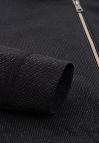 Schwarzer, texturierter Stoff mit kleinen, erhabenen Mustern, ausgestattet mit einem Reißverschluss und aufgerolltem Bund. Das Design betont eine glatte, gleichmäßige Oberfläche.