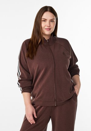 Kvinde med langt brunt hår iført en brun træningsdragt med lynlås og hvide striber på ærmerne, hænderne i lommerne, let smilende.