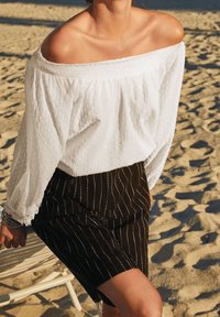 Blouse blanche à épaules dénudées avec des détails texturés, associée à une jupe noire à rayures. Le fond sableux ajoute du contraste.