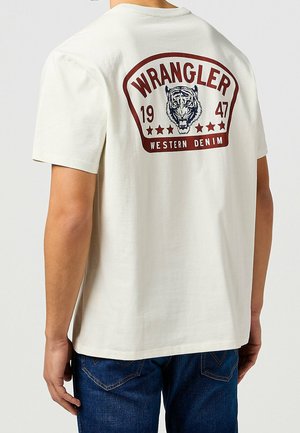 Osoba ubrana w białą koszulkę Wrangler z grafiką tygrysa, napisem "1947" oraz "Western Denim" na plecach, w połączeniu z niebieskimi dżinsami.