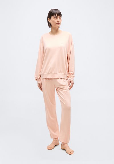 Γυναίκα όρθια, ξυπόλυτη, φορώντας ανοιχτό ροζ φαρδύ φούτερ και matching παντελόνι, σε απλό φωτεινό φόντο.