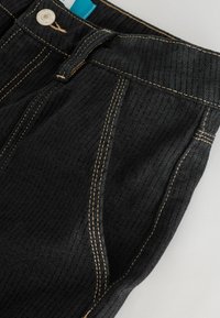 Czarne spodnie dżinsowe z teksturowanym materiałem, kontrastującym żółtym przeszyciem, metalowym zapięciem oraz niebieską metką wewnątrz w pobliżu pasa.