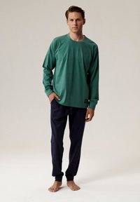 Zielona koszula z długim rękawem, okrągłym dekoltem i ma�łym logo, w połączeniu z ciemnoniebieskimi spodniami. Oba elementy charakteryzują się gładką, miękką teksturą.