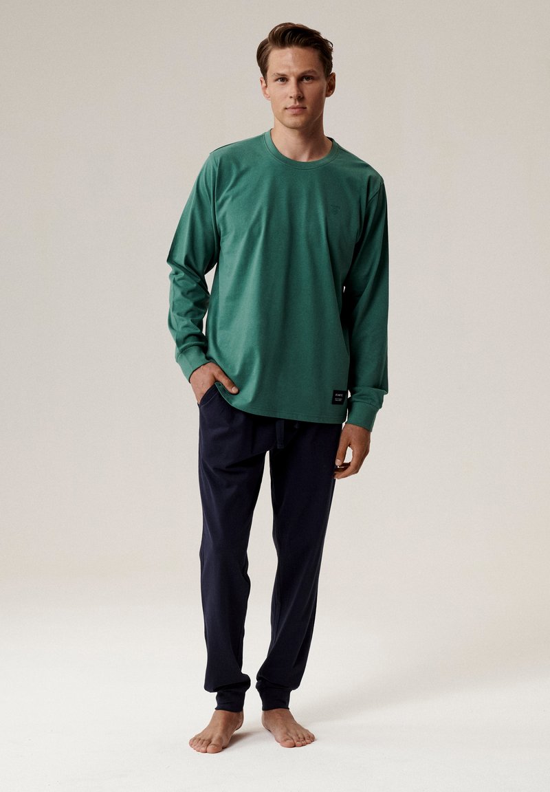 Zielona koszula z długim rękawem, okrągłym dekoltem i małym logo, w połączeniu z ciemnoniebieskimi spodniami. Oba elementy charakteryzują się gładką, miękką teksturą.