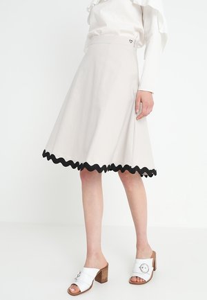 Kobieta ubrana w kremową spódnicę w kształcie litery A z czarnym falowanym brzegiem, białą bluzkę z falbankami oraz białe sandały na obcasie z drewnianym słupkiem.