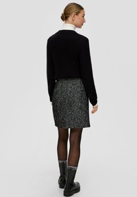Un pull noir à côtes associé à une jupe en tweed grise. Le modèle porte des bottes noires avec des détails texturés. Fond uni.