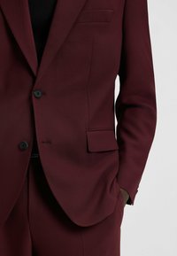 Burgundy suit jacket with a smooth texture, single-breasted design, two black buttons, and a streamlined fit; features a welt pocket.
