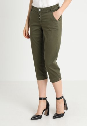 Femme portant un pantalon court vert olive avec braguette à boutons, poches avant et talons à brides chevilles noirs, debout sur un sol blanc.