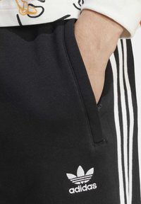Fatos de treino pretos com design de três riscas brancas e pequeno logo branco da Adidas na bolsa, feitos de um material suave. Apresenta um bolso lateral.