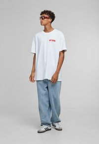 White oversized t-shirt with red "DAYS BEFORE" text, paired with light blue baggy jeans and grey-black-striped sneakers, complemented by red sunglasses.