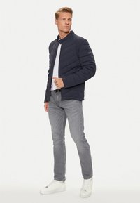 Giacca a vento blu navy con colletto alto e motivo a zigzag, abbinata a jeans grigi e scarpe da ginnastica bianche. Materiale liscio e lucido.