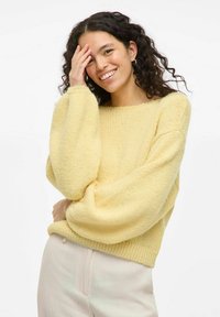 Svetlo rumena, pletena pulover s širokim čolnom izrezom in obsežnimi rokavi. Prepleten rob in manšete, mehak material. V kombinaciji z svetlimi hlačami.