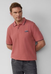 Man in een zalmroze polo met een klein autologo op de borst, gecombineerd met donkergrijze jeans, staande tegen een effen muur.