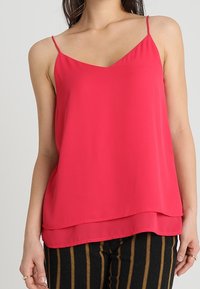 Woman wearing a sleeveless bright pink layered camisole top and black pants with vertical brown stripes, standing against a plain background.