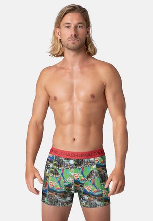 Man zonder shirt met schouderlang blond haar, gekleed in kleurrijke, patroonrijke boxershorts met een rode tailleband waarop "MUCHACHOMALO" staat.