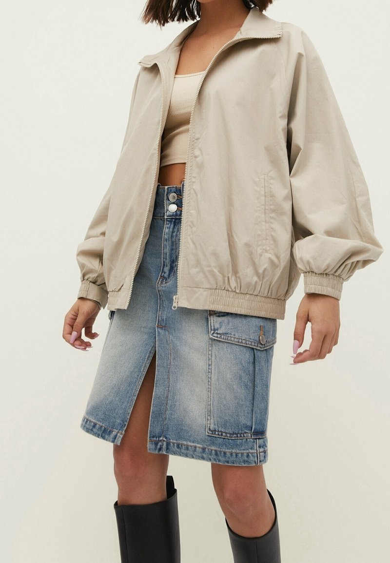 Femme portant une veste beige zippée, un crop top beige, une jupe en jean bleue comportant une fente avant et une poche, ainsi que des bottes noires montant jusqu'aux genoux.