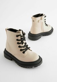 Stivali alla caviglia in similpelle beige con lacci neri, suola in gomma nera texturizzata, cerniera laterale e un accento a forma di stella sul tallone.