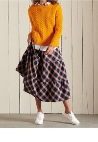 Oranger Kabelstrickpullover, über einem weißen Hemd getragen, kombiniert mit einem navyblauen, karierter, gestufter Rock. Dazu weiße Sneaker.
