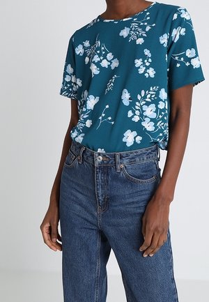 Person wearing a teal short-sleeve shirt with white floral print tucked into high-waisted blue denim jeans.
