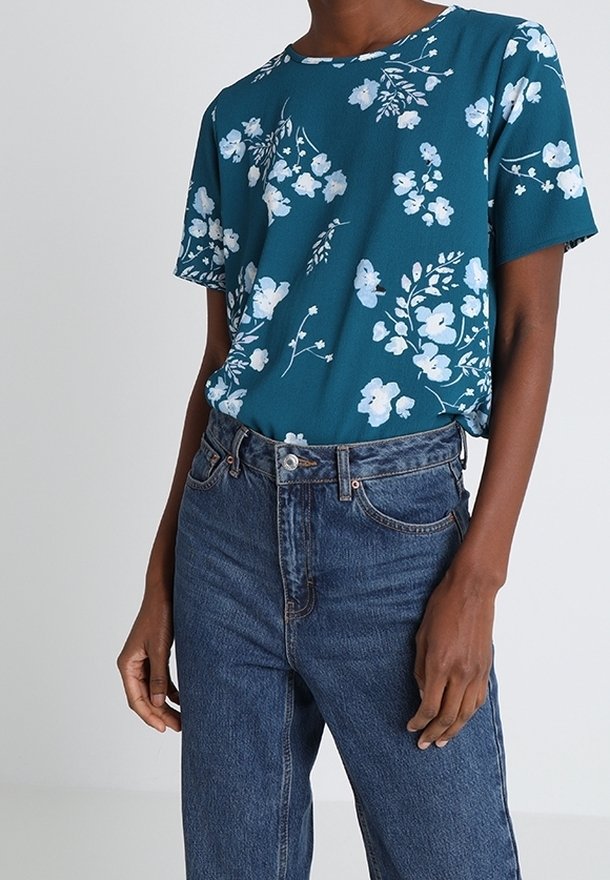 Personne portant une chemise à manches courtes bleu sarcelle avec un imprimé floral blanc, rentrée dans un jean en denim bleu taille haute.