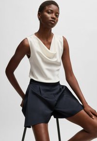 Ärmelloses Oberteil mit Schalkragen in Creme, kombiniert mit hochgeschnittenen, gefalteten navyblauen Shorts. Der glatte Stoff und die entspannte Passform schaffen eine moderne Silhouette.