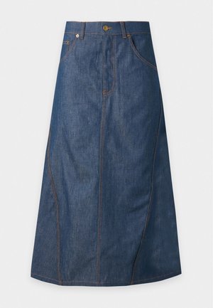 Femme portant un pull blanc Moschino avec un motif ourson, une longue jupe en jean bleue et des sandales noires à talons, debout avec une main dans la poche.