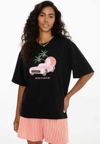 Žena s dlouhými kudrnatými vlasy nosí černé tričko s růžovým autem, palmami a textem "Příliš skvělá na to, abych stála ve frontě", doplněné oranžovo pruhovanými šortkami.