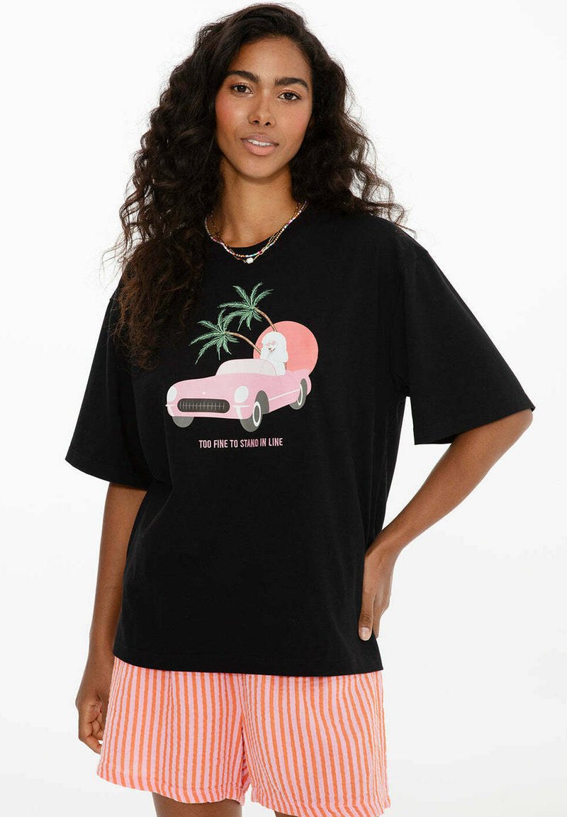 Žena s dlouhými kudrnatými vlasy nosí černé tričko s růžovým autem, palmami a textem "Příliš skvělá na to, abych stála ve frontě", doplněné oranžovo pruhovanými šortkami.
