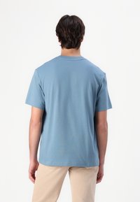 Homme debout de dos, portant un t-shirt à manches courtes bleu clair uni et un pantalon beige, sur un fond blanc.