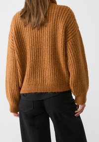 Personne portant un pull marron texturé en maille avec des manches longues et un pantalon noir, vue de dos sur un fond uni.