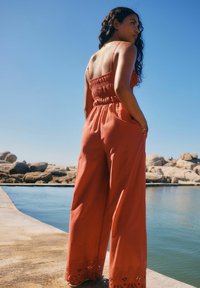 Romper en terracotta avec un corsage smocké, jambes larges et accents brodés sur les ourlets, présentant des bretelles spaghetti et des poches.