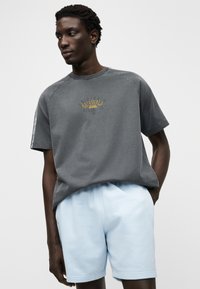 Szürke pamut póló sárga grafikával a közepén, világoskék rövidnadrággal párosítva. Az anyag sima tapintású és laza szabású.