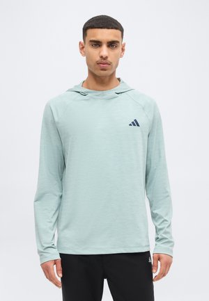 Νέος άνδρας που φοράει αθλητική μπλούζα με κουκούλα και μακριά μανίκια σε ανοιχτό πράσινο χρώμα, με μικρό σκούρο λογότυπο στο στήθος και μαύρο παντελόνι, στέκεται μπροστά σε λευκό φόντο.