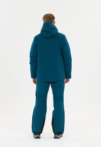 Person steht mit dem Rücken zur Kamera, trägt eine türkisfarbene Kapuzenjacke und passende Hose, mit grauen und schwarzen Stiefeln vor einem schlichten weißen Hintergrund.