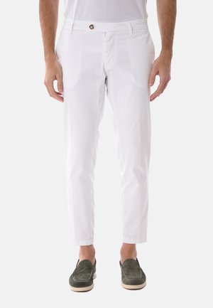 Uomo con pantaloni bianchi a taglio slim e mocassini in camoscio verde, in piedi contro uno sfondo semplice chiaro.