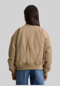 Beige Bomberjacke mit geripptem Kragen und Bündchen, lockerer Schnitt und glatter Textur. Über einer blauen Jeans getragen, von hinten betrachtet.