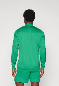 Πράσινο αθλητικό μπουφάν με ψηλό γιακά, μακριά μανίκια και λευκές ρίγες στα πλάγια. Κατασκευασμένο από λείο, ελαφρύ ύφασμα, συνοδευόμενο από ταιριαστά σορτς.