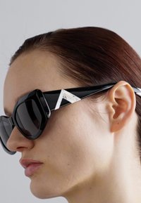 Black oversized sunglasses with angular shapes, featuring a white accent on the temple and dark lenses, highlighting a bold design.