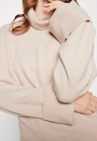 Beżowy sweter z golfem o miękkiej teksturze, z szerokimi ściągaczami i luźnym krojem, sięgającym do talii.
