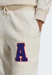 Närbild av beige sweatpants med ett blått och rött bokstavs-patch "A" och tre vertikala ränder, hand i framfickan.