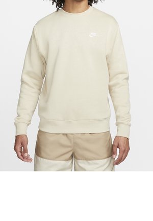 Man wearing beige Nike crewneck sweatshirt and two-tone beige shorts, standing with arms relaxed at sides.