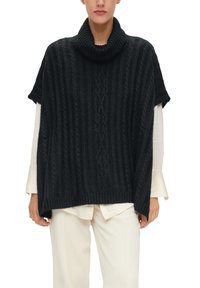 Zwarte gebreide poncho met een hoge kraag, geribbelde textuur en korte mouwen, gelaagd over een lichtgekleurde blouse en wijde broek.