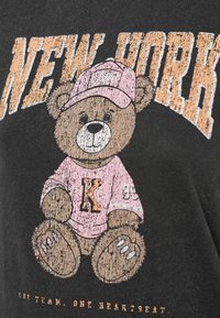 Γκρι μπλουζάκι με γραφικό ενός καφέ αρκούδου που φοράει ροζ πουκάμισο και καπέλο, με το κείμενο «NEW YORK» πάνω και «ONE HEARTBEAT» κάτω.