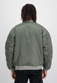 Jachetă tip bomber de culoare verde măslinie, dintr-un material lucios, cu manșete și tiv accesorizate cu nervuri și o croială lejeră. Prezintă umăruri căptușite și o buzunare cu fermoar pe mânecă.
