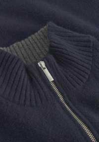 Marineblauer Wollpullover mit einem gerippten grauen Kragen, der einen silbernen Reißverschluss aufweist. Die Textur wirkt weich, mit einem feinen Strickmuster am Hals.